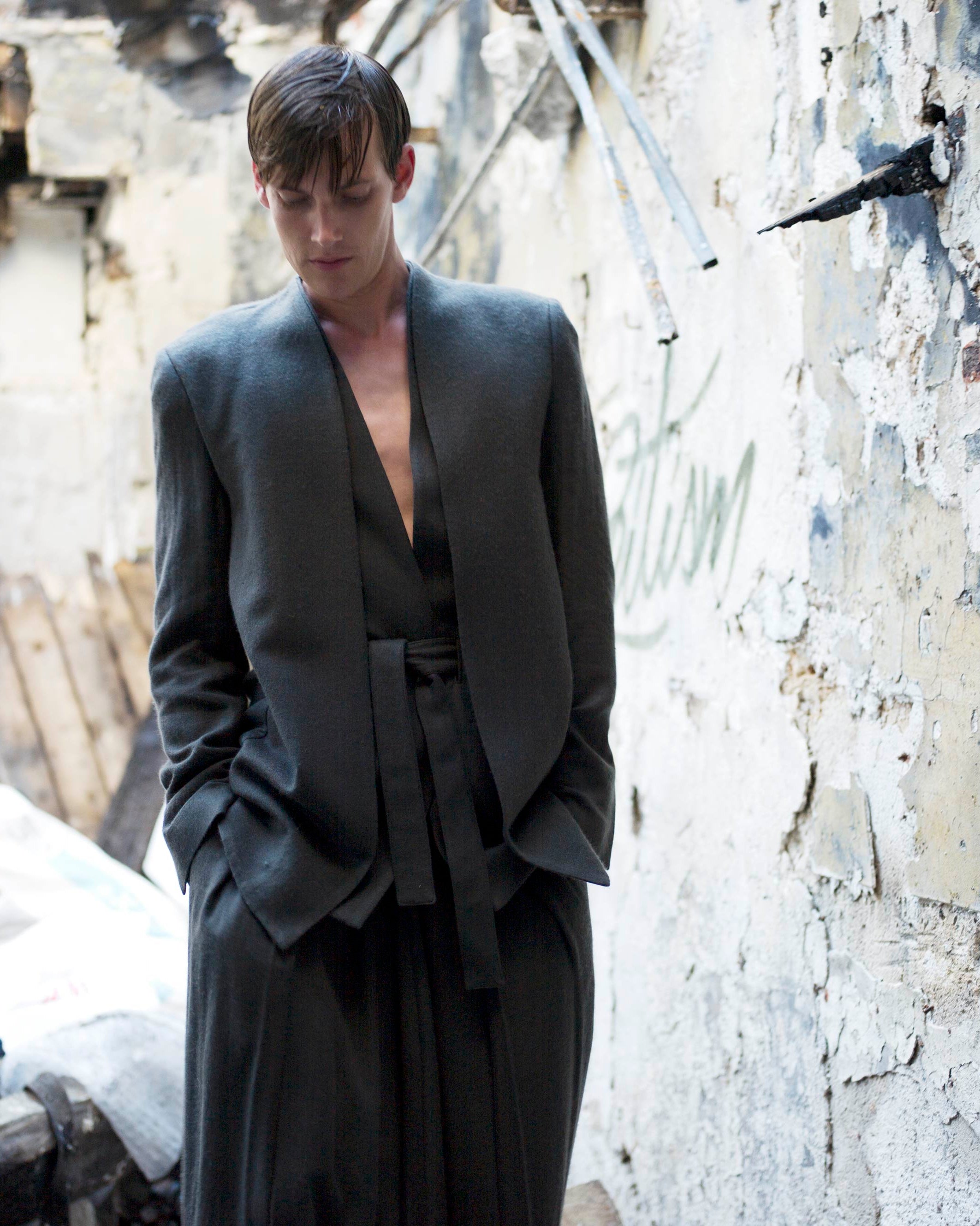A person in a dark, loose-fitting outfit stands in an abandoned, dilapidated building with peeling walls and rubble. They have a contemplative expression, hands in pockets, and their hair is slightly wet. The scene conveys a somber, reflective mood.
