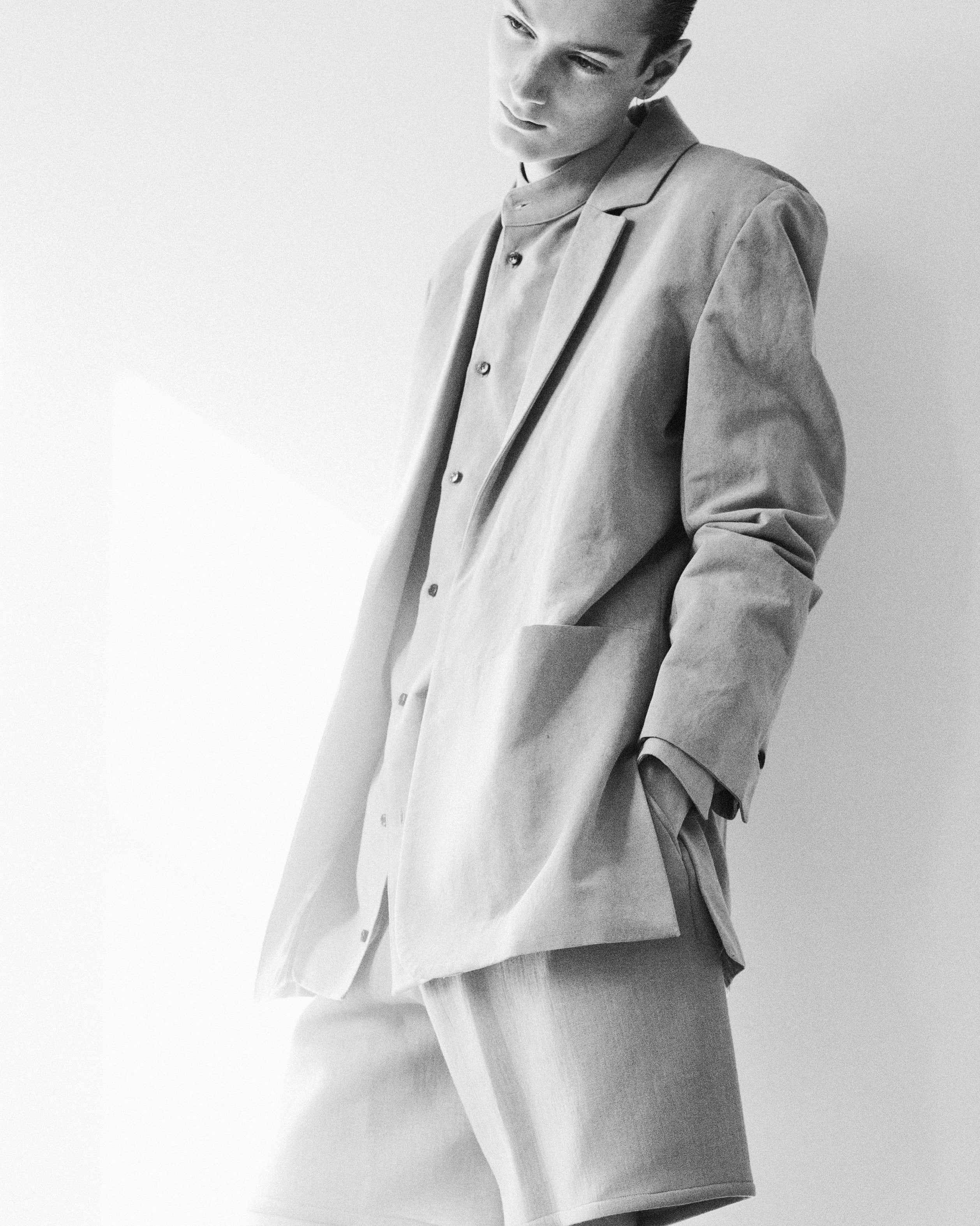 A person in grayscale wearing a loose-fitting suit and shorts leans against a wall. The soft lighting creates a relaxed and stylish atmosphere.
