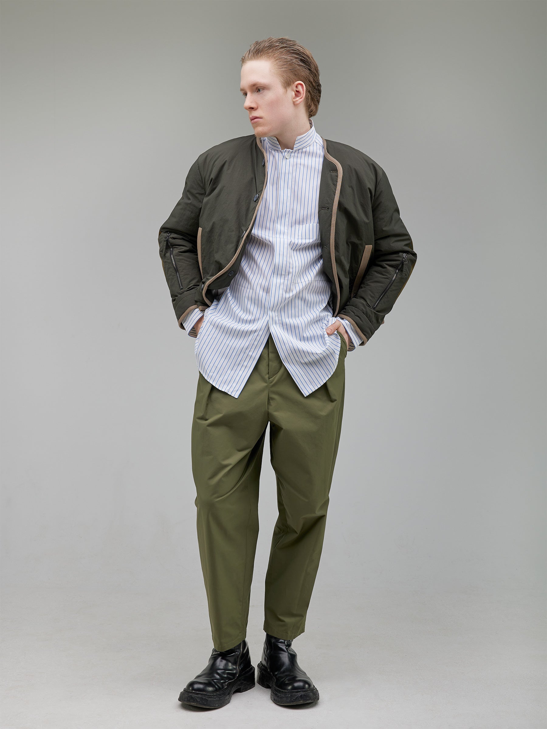 Insulated liner with mesh elbow panels, taped piping, and vintage horn button closure. Functional zip pockets at the lower arms add an adaptive touch.
Shirt: Oversized mandarin-collar shirt in white with light blue stripes. A hidden placket ensures a clean, uninterrupted finish.
Fabric: 100% Cotton
Buttons: White Trocas Mother of Pearl
Bottom: Tailored rounded trousers with a structured waist and precise pleating.