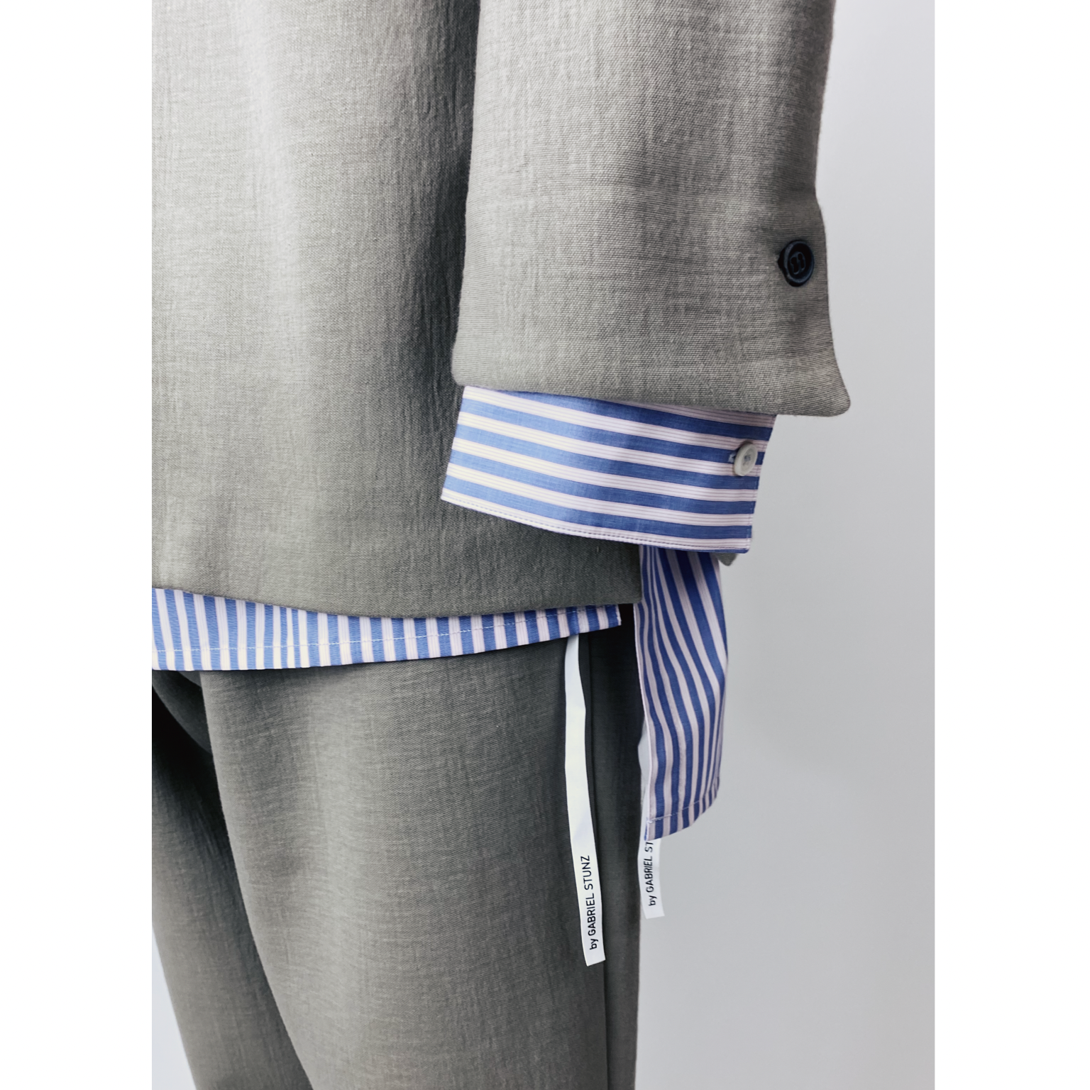 Close-up of a person wearing a gray suit with a blue and white striped shirt underneath. The shirt cuffs extend past the suit sleeves, displaying white tags with text. The suit features a black button detail on the sleeve.