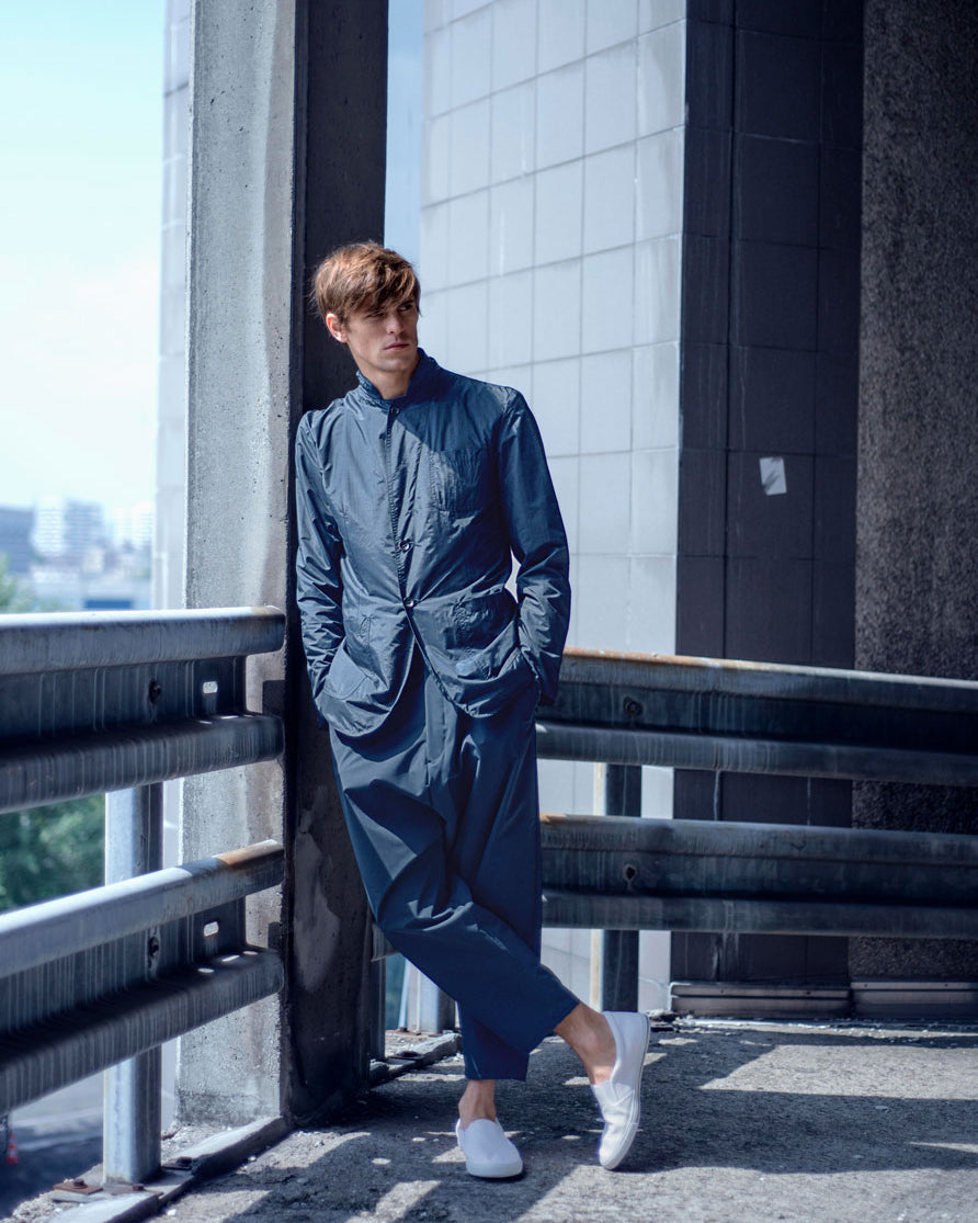 A person in a blue outfit stands casually against a railing in an urban setting. They are wearing white slip-on shoes. The background features concrete walls and a distant view of the city skyline.