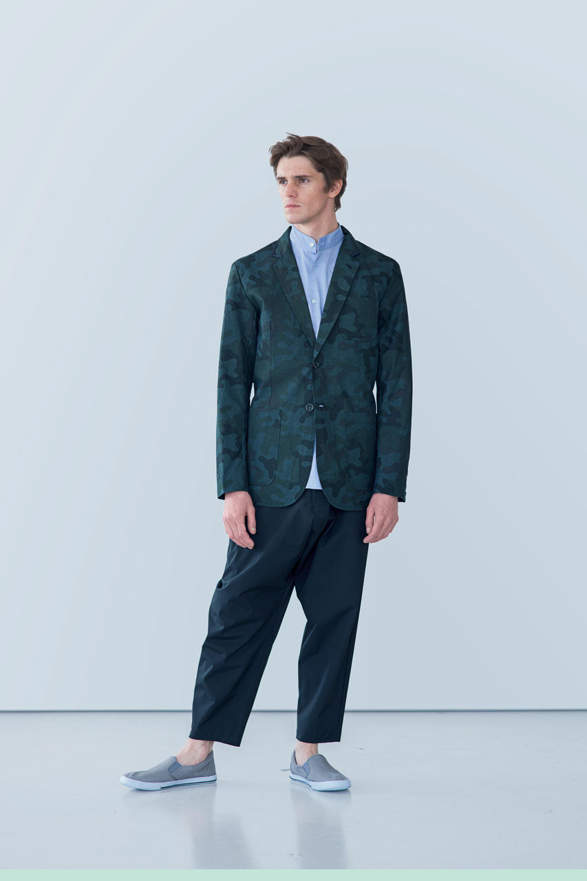 A man stands in a minimalistic setting wearing the gabrielstunz POCKETPARACHUTE® Camouflage Jacket crafted from performance fabric over a light blue shirt and dark cropped trousers. He has gray slip-on shoes. The plain background enhances the modern, stylish look.