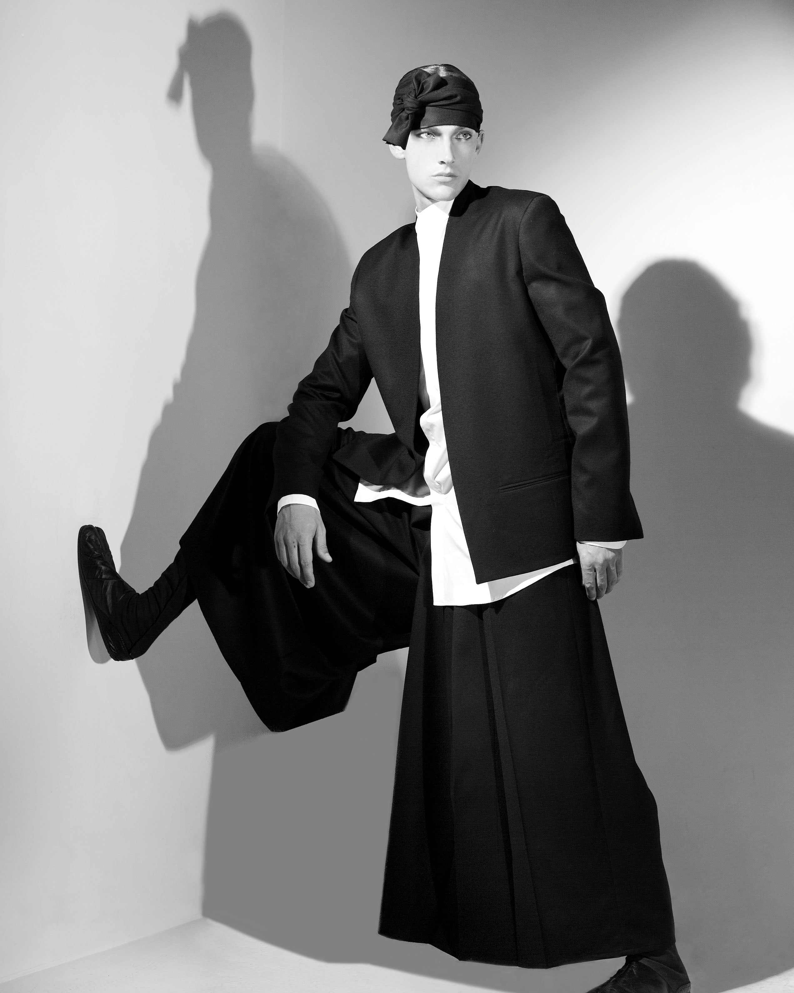A person in a monochrome outfit poses dramatically against a wall. They wear a black jacket, long black skirt, white shirt, and black cap. One leg is bent with the foot resting on the wall, casting a shadow in the soft lighting.
