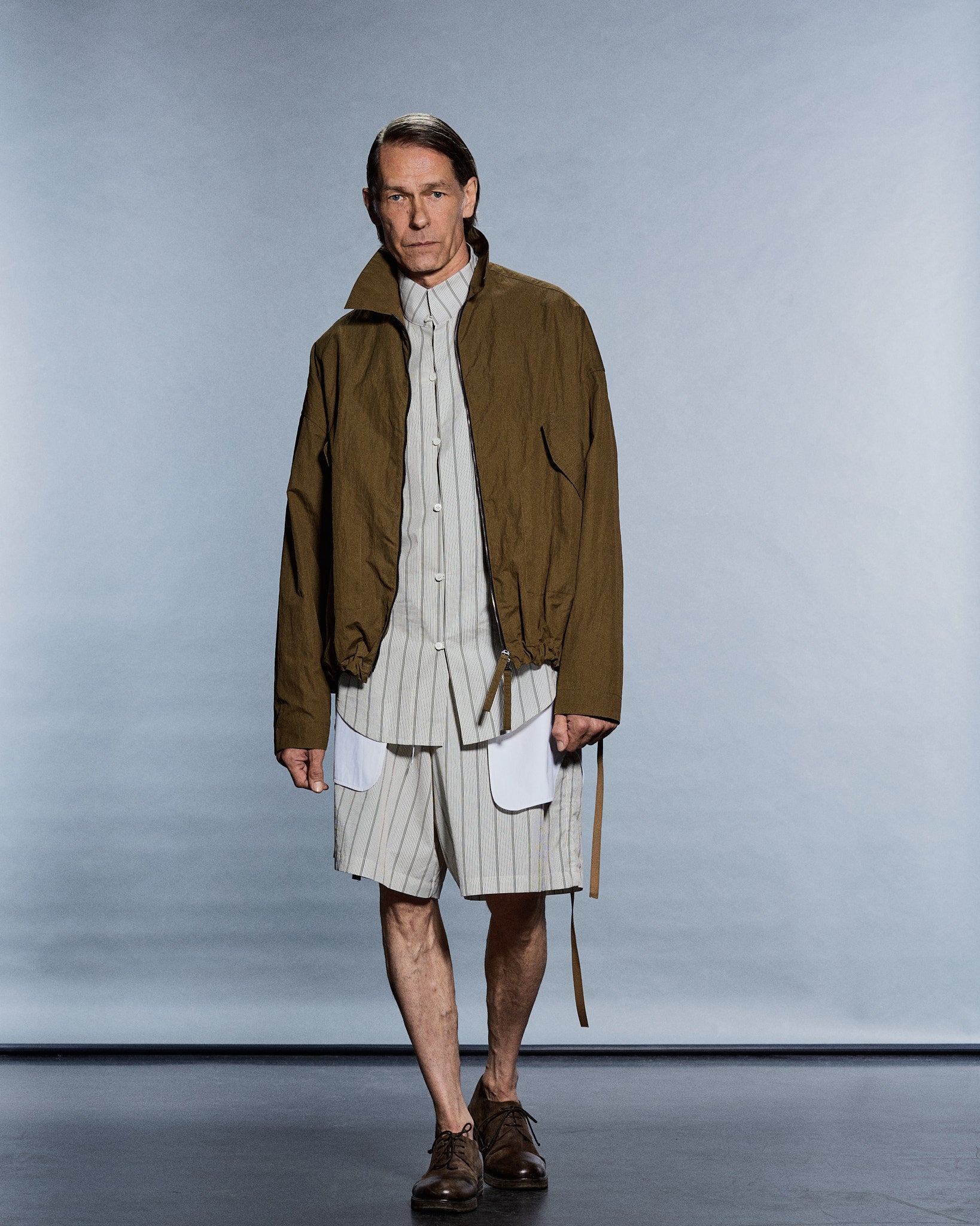 Model in an olive zip blouson worn open over a striped shirt and shorts — GABRIELSTUNZ APLOMB SS27.