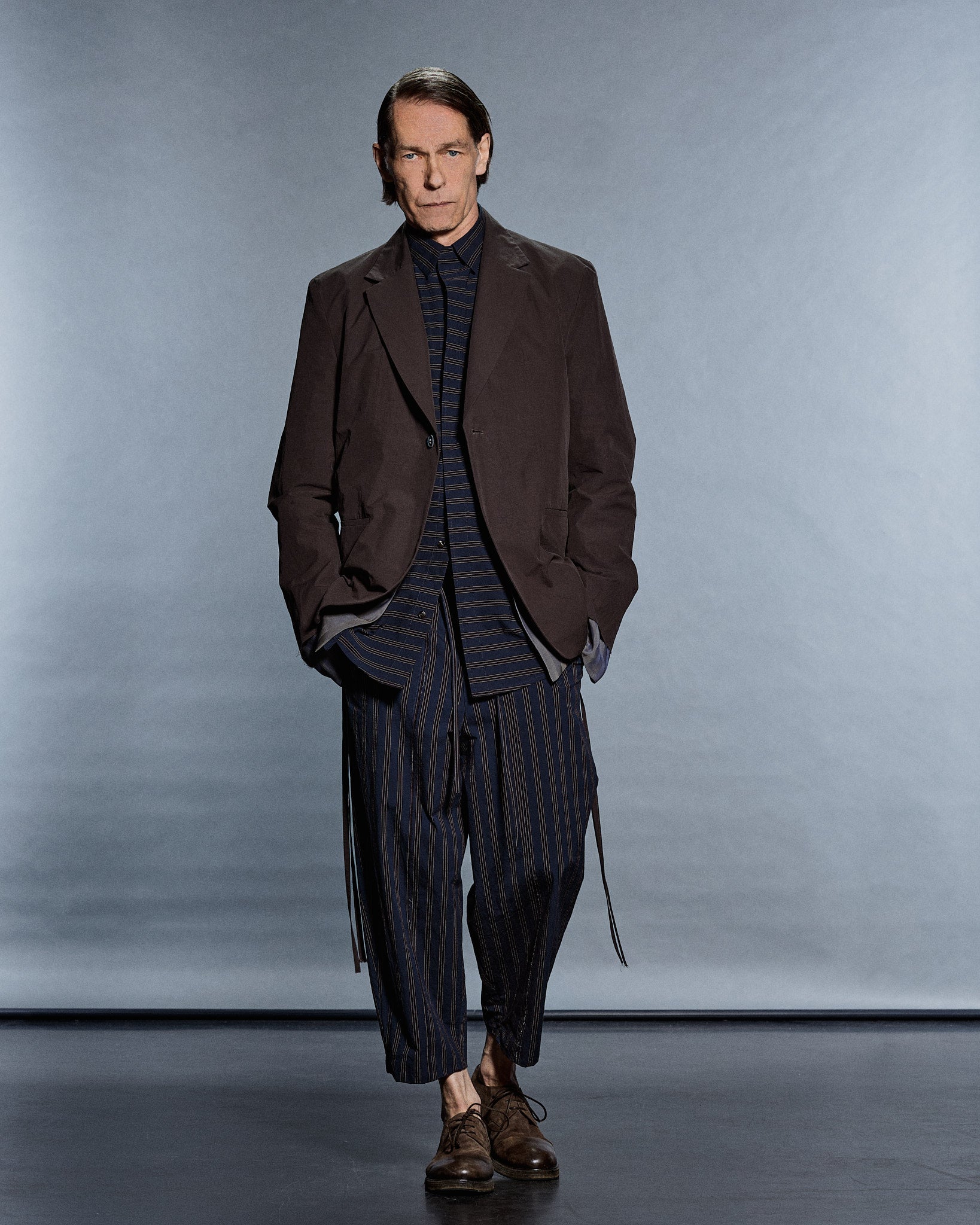 Full-length brown tailored blazer worn open over a navy fine-stripe shirt and matching cropped trousers with trailing ties — GABRIELSTUNZ APLOMB SS27