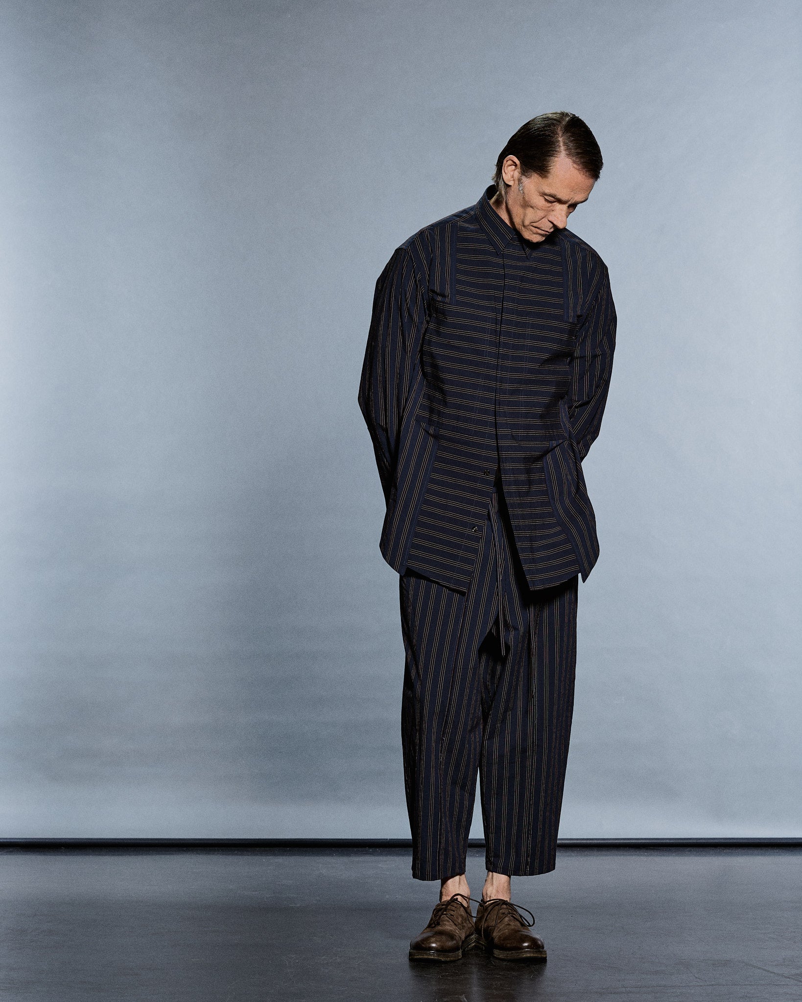 Full-length navy-and-tan pinstripe shirt-jacket and tapered trousers, head bowed — GABRIELSTUNZ APLOMB SS27.
