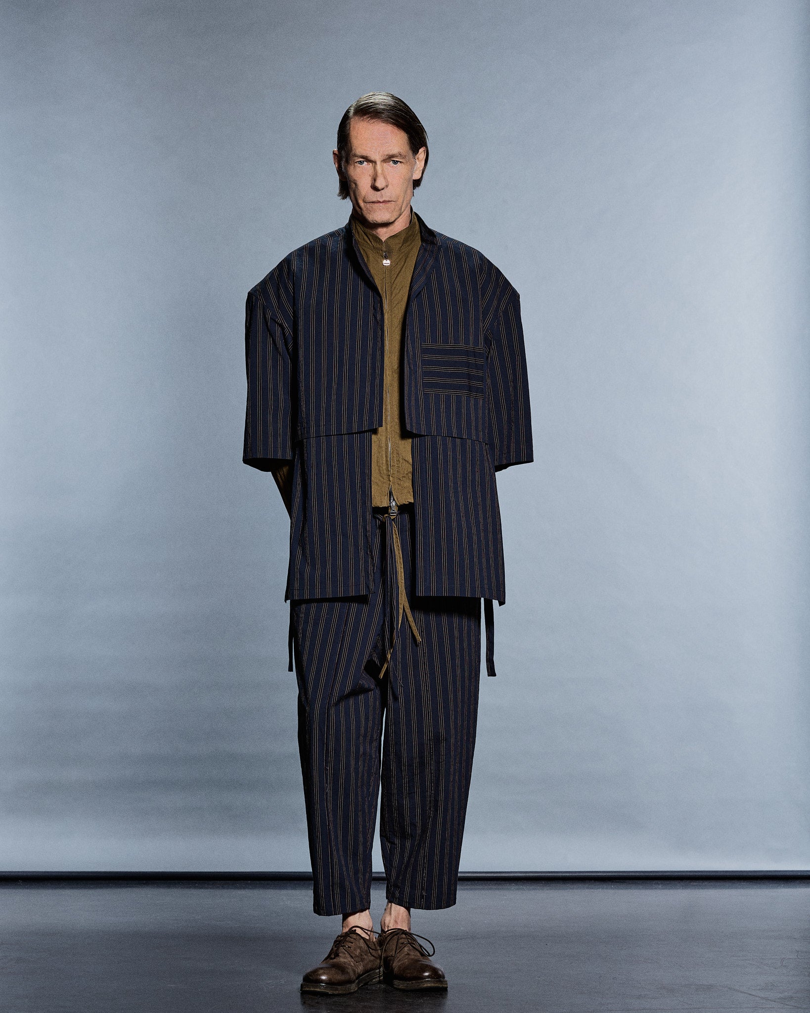 Cropped navy-stripe overlayer over an olive zip shirt and matching trousers — GABRIELSTUNZ APLOMB SS27.