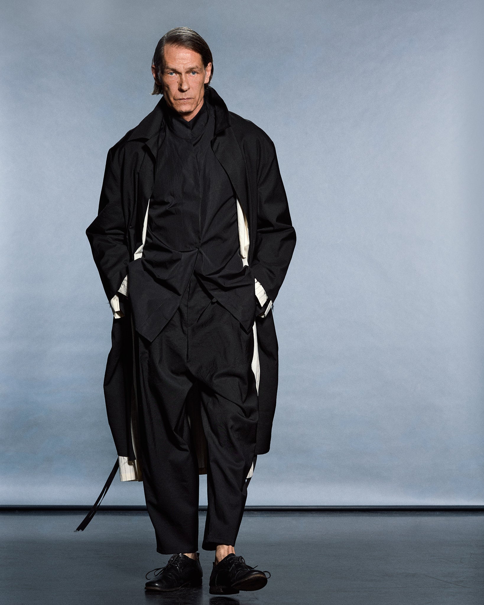 Full-length layered black look — draped overshirt, scarf-collar and cropped trousers over pinstripe lining — GABRIELSTUNZ APLOMB SS27.