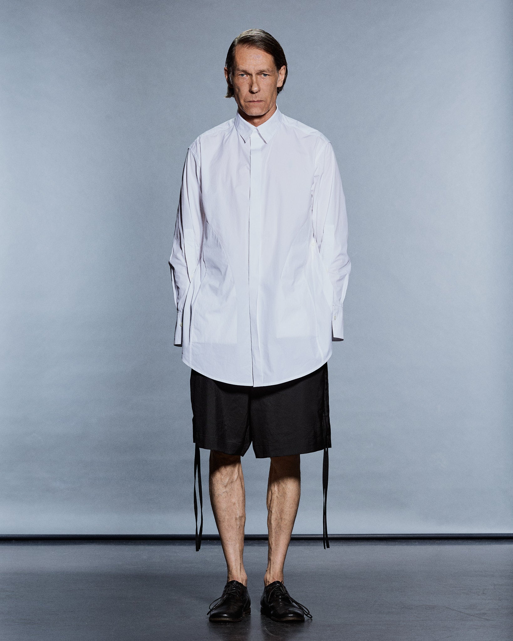 Full-length oversized white cotton shirt worn over black shorts with side ties — GABRIELSTUNZ APLOMB SS27.